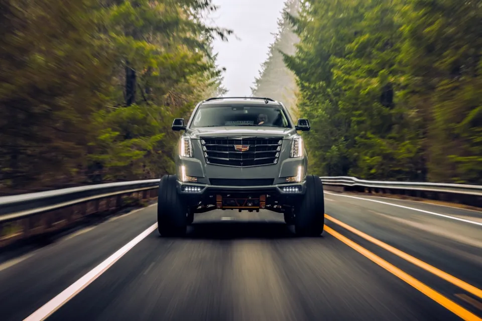 Cadillac Escalade SUV driving toward camera on black asphalt road surrounded by forest